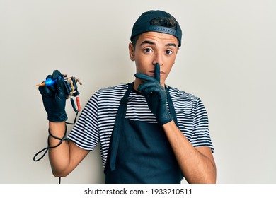 Young handsome african american man tattoo artist wearing professional uniform and gloves holding tattooer machine asking to be quiet with finger on lips. silence and secret concept. 