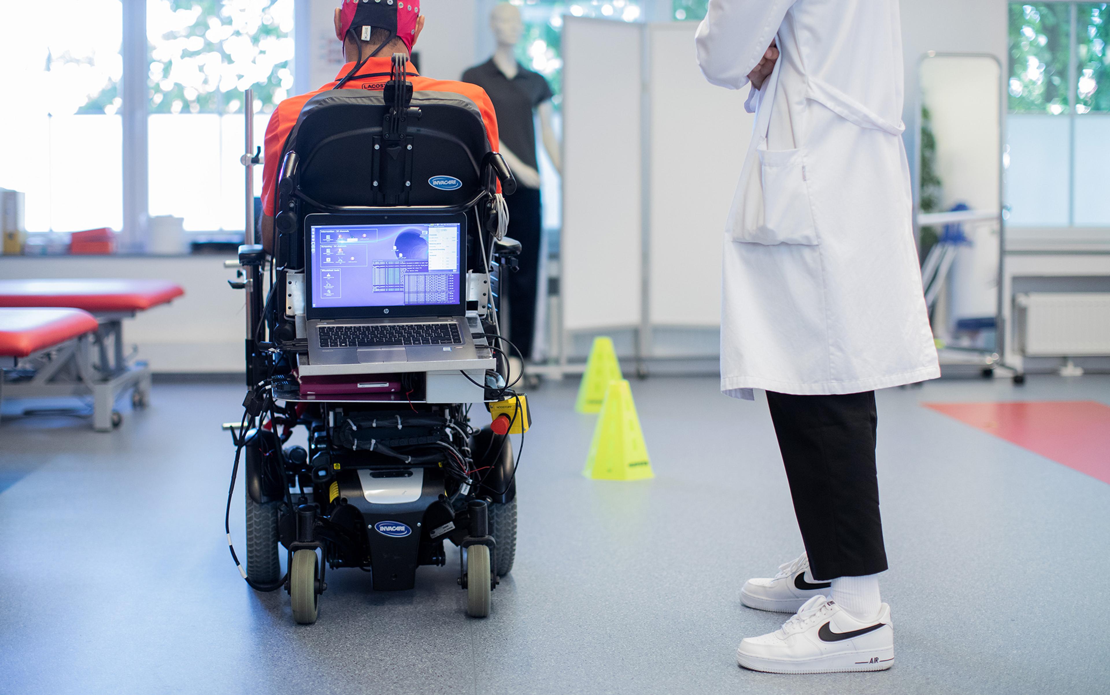 Person in a wheelchair with a laptop, wearing a monitoring cap, and a doctor in a lab coat standing nearby in a clinical setting.