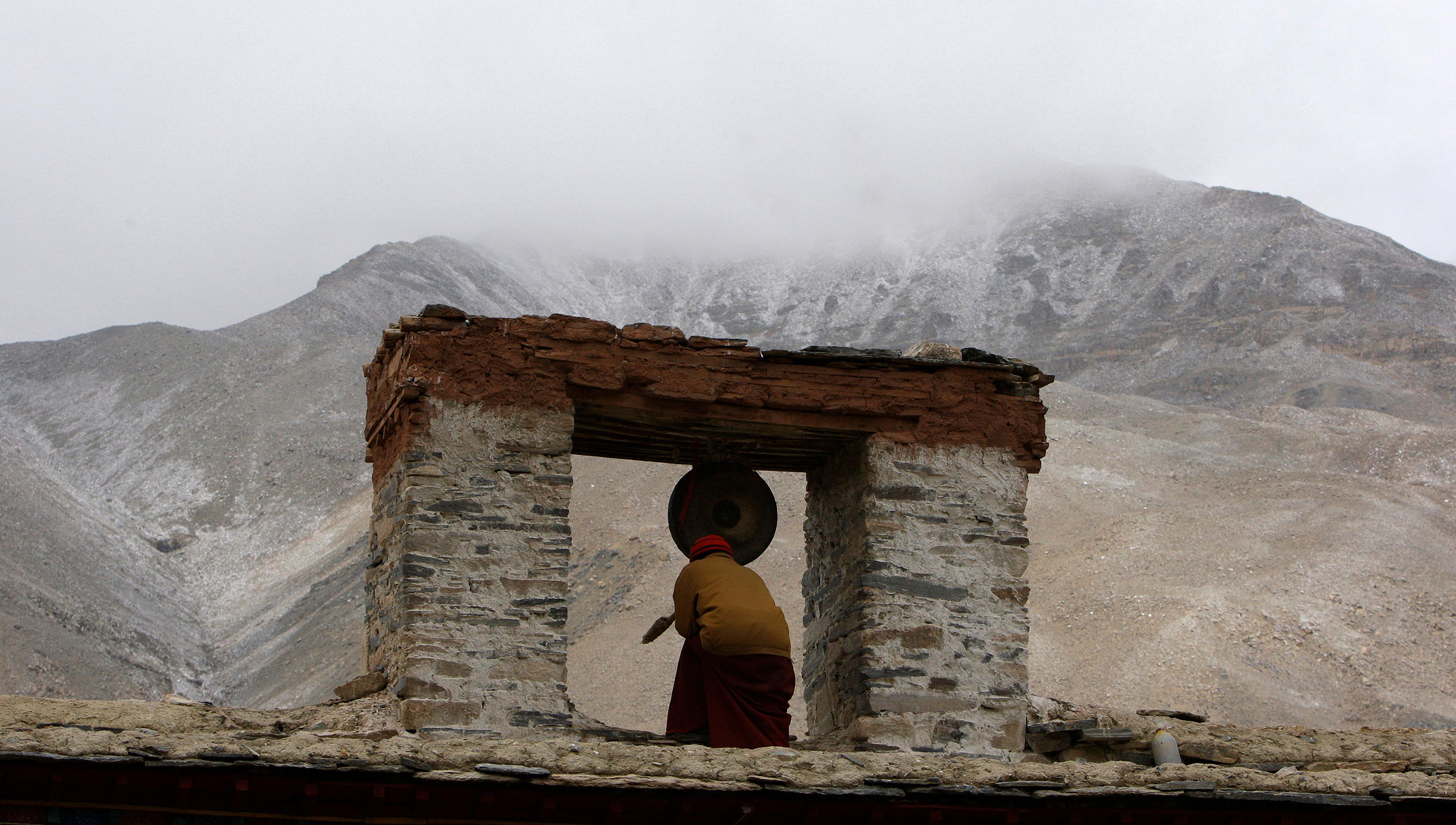 A person kneeling in front of a gong in an ancient stone structure, with a misty mountain landscape in the background.