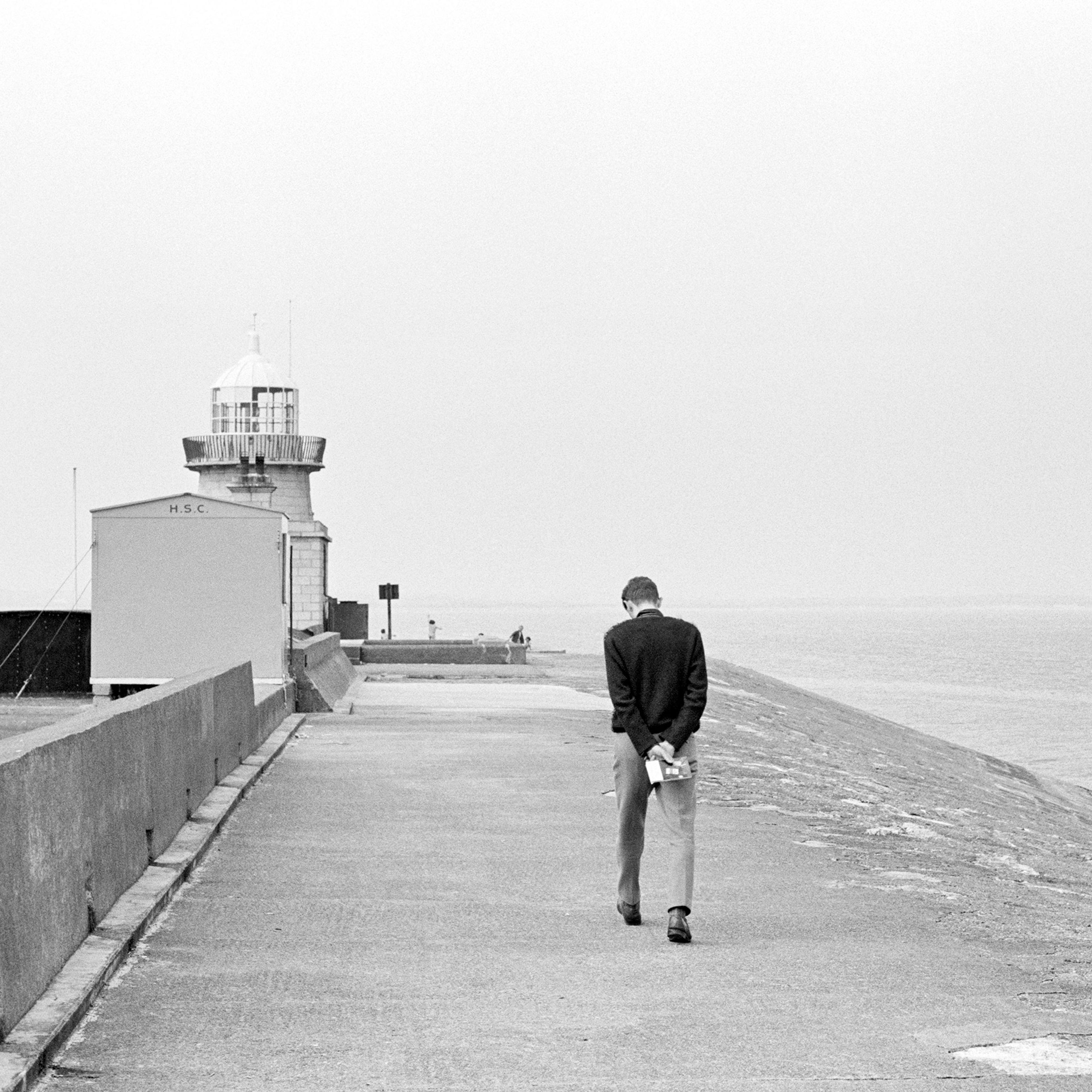 A person walking on a pier towards a lighthouse, holding a book behind their back, with the sea on the right under a grey sky.