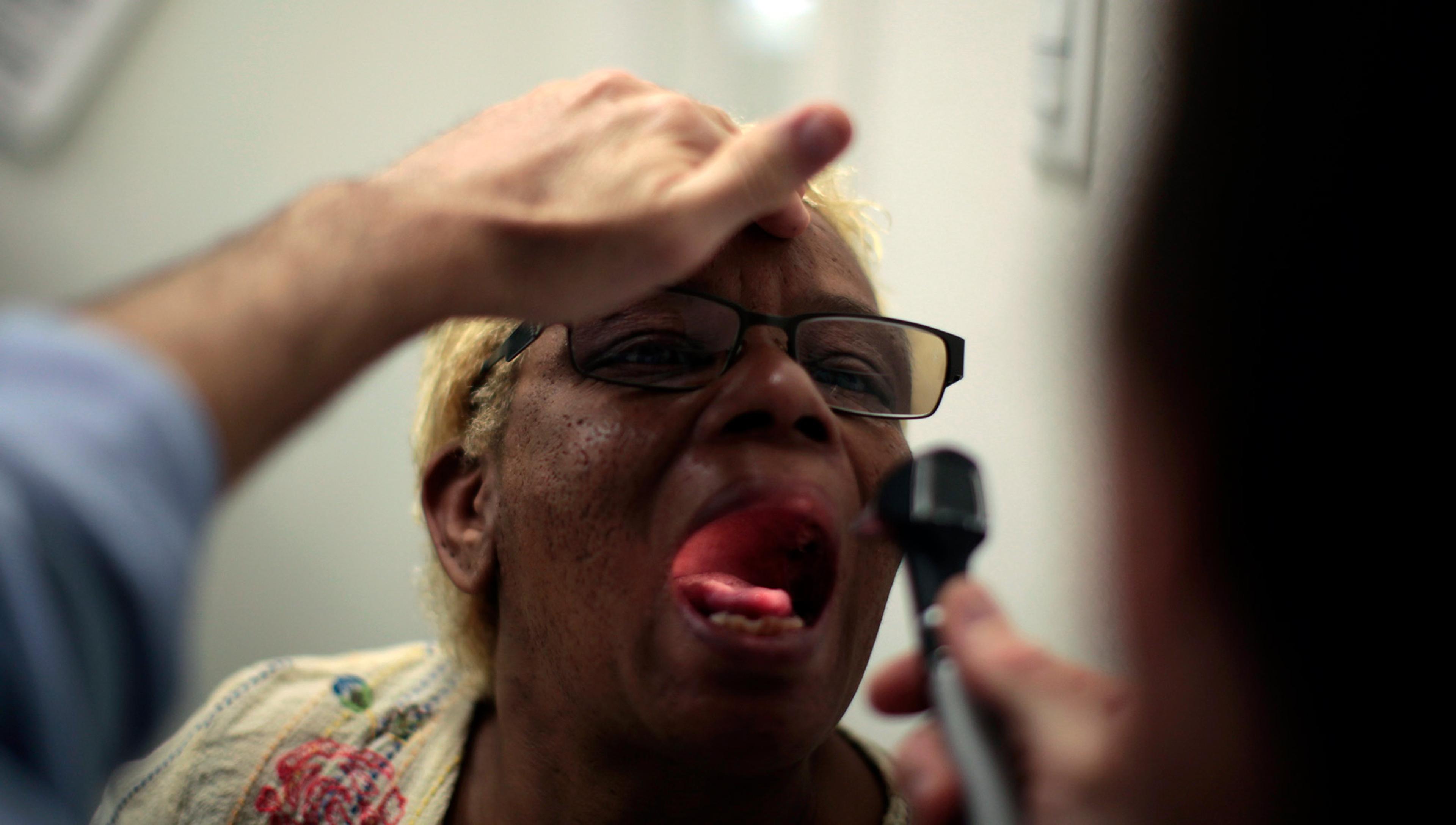 A doctor examines a patient’s mouth using a small light. The patient is wearing glasses and has their mouth open wide.