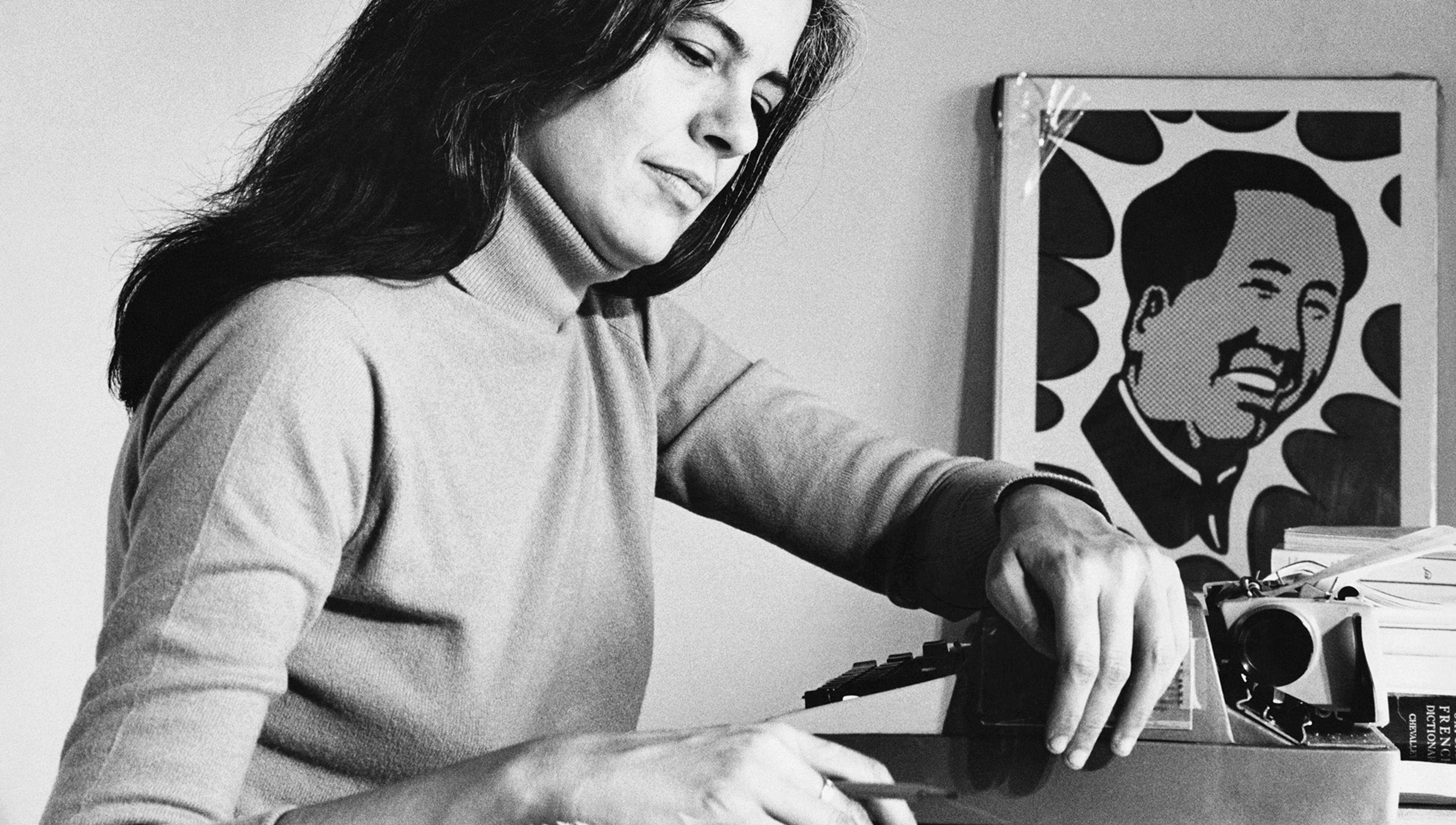 Woman with long dark hair sitting at a desk, using a typewriter. Art of a smiling face in the background.