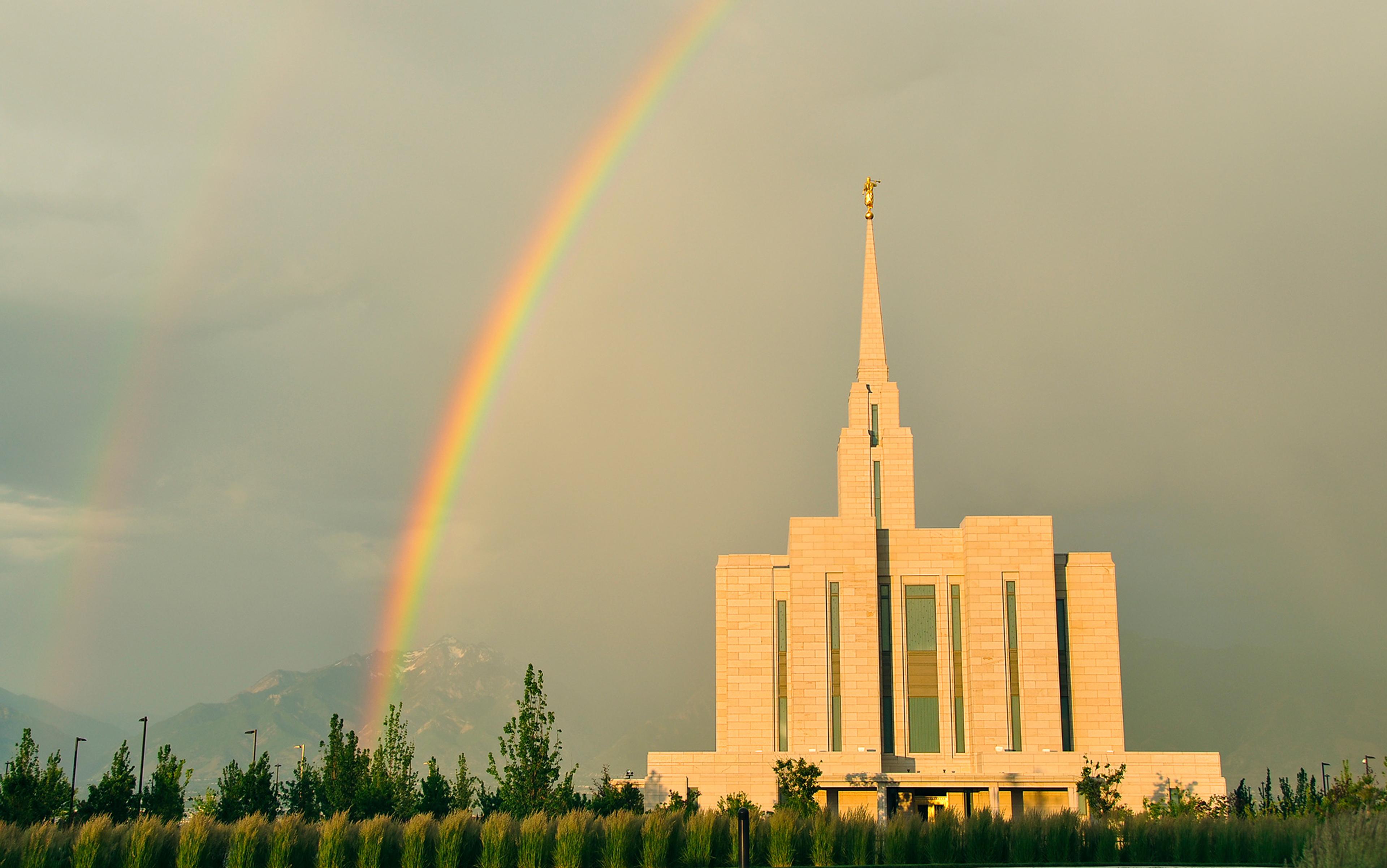 Photo of a white temple with modern design features, a golden statue on top and a bright double rainbow in the cloudy sky.