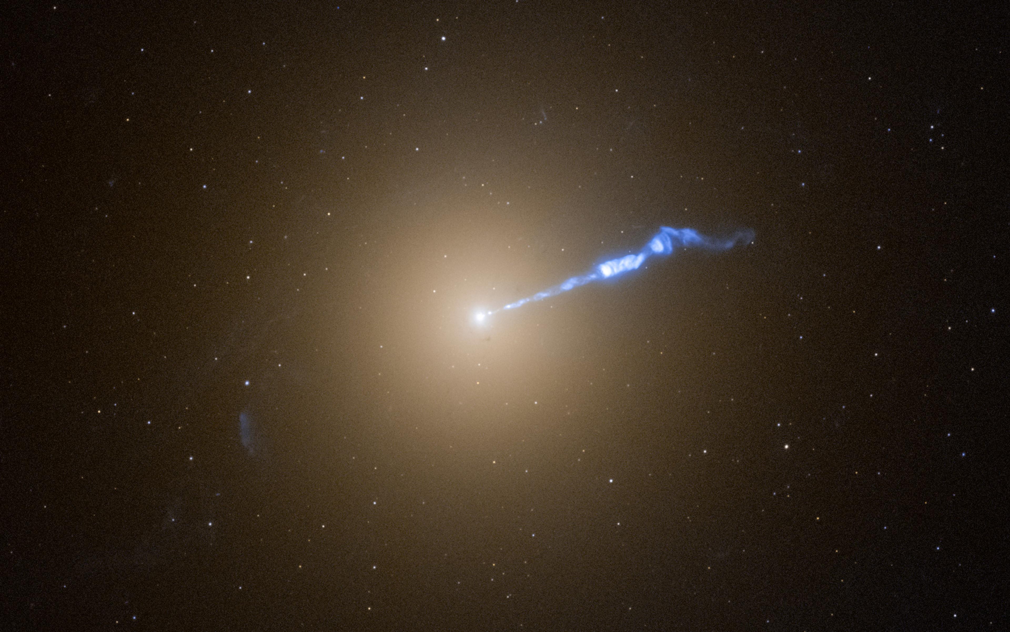 Photo of a galaxy with a bright centre emitting a blue jet of energy against a background of numerous stars in space.