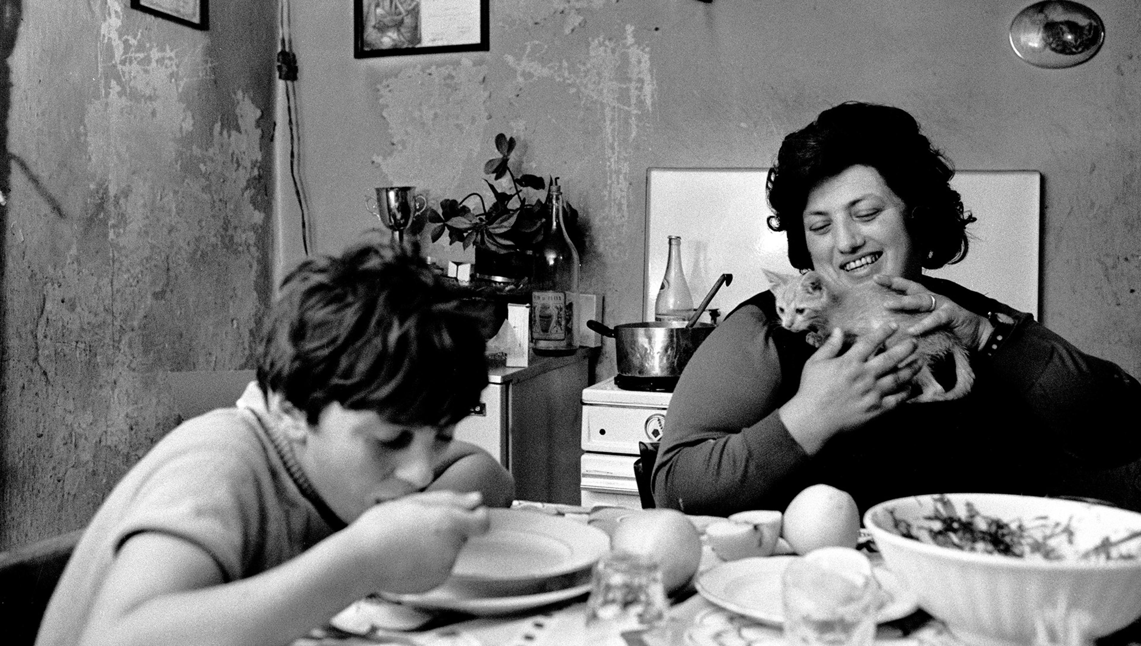 A boy eats at a table while a smiling woman holds a kitten in a rustic kitchen with peeling walls and various items on a counter.