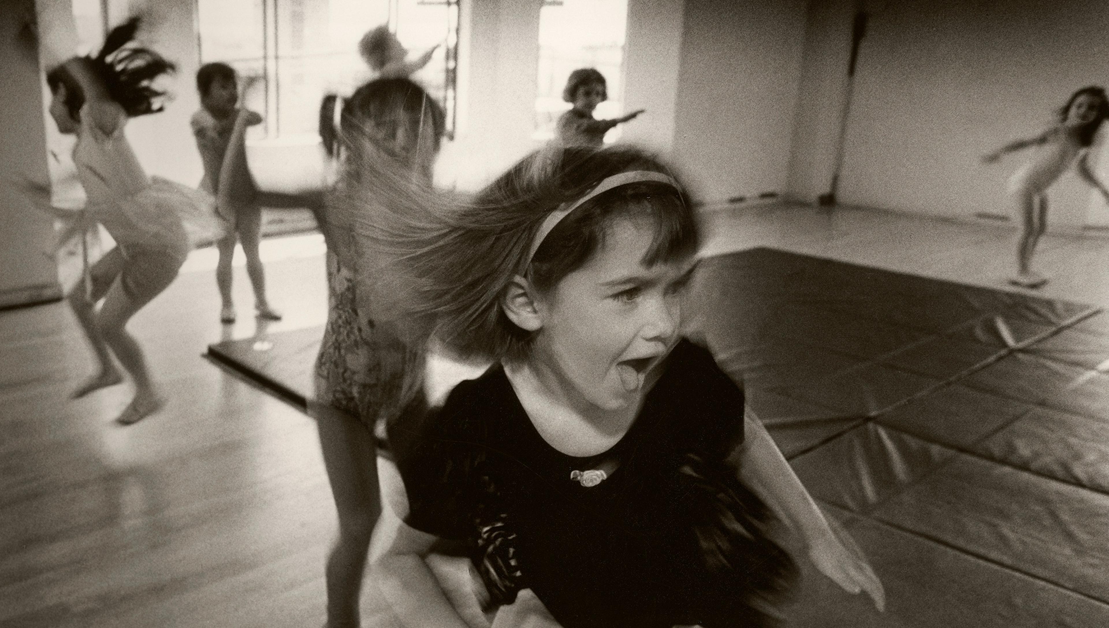 Black-and-white photo of young children dancing in a studio, with one girl in the foreground, mouth open and hair swinging.