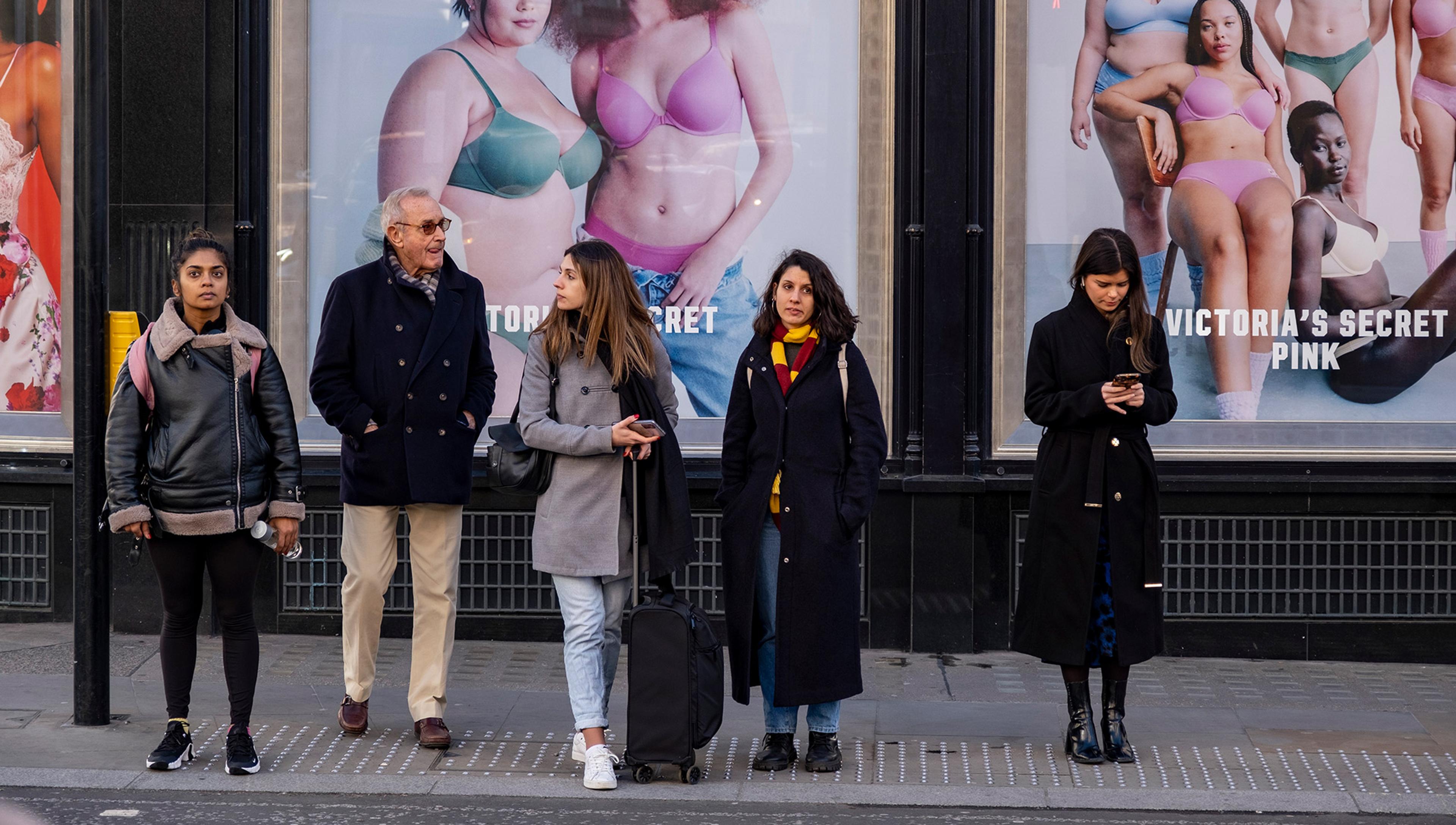 Five people wait to cross by Victoria’s Secret posters; some use their phones, while others look around.