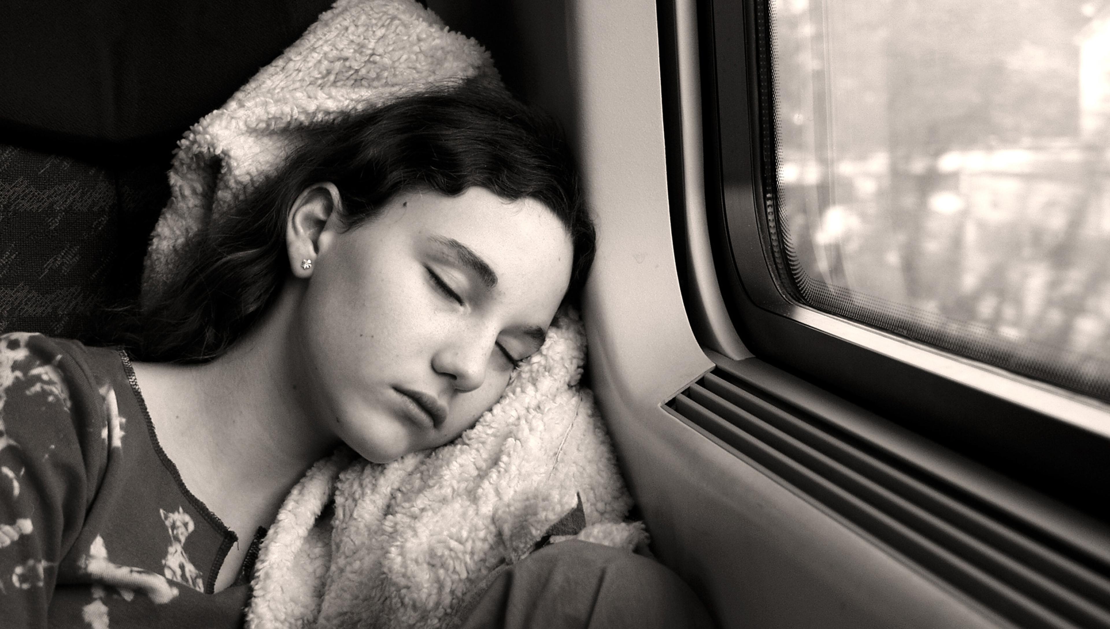 Black-and-white photo of a person sleeping against a window on a train, head resting on a pillow, outside view blurred.