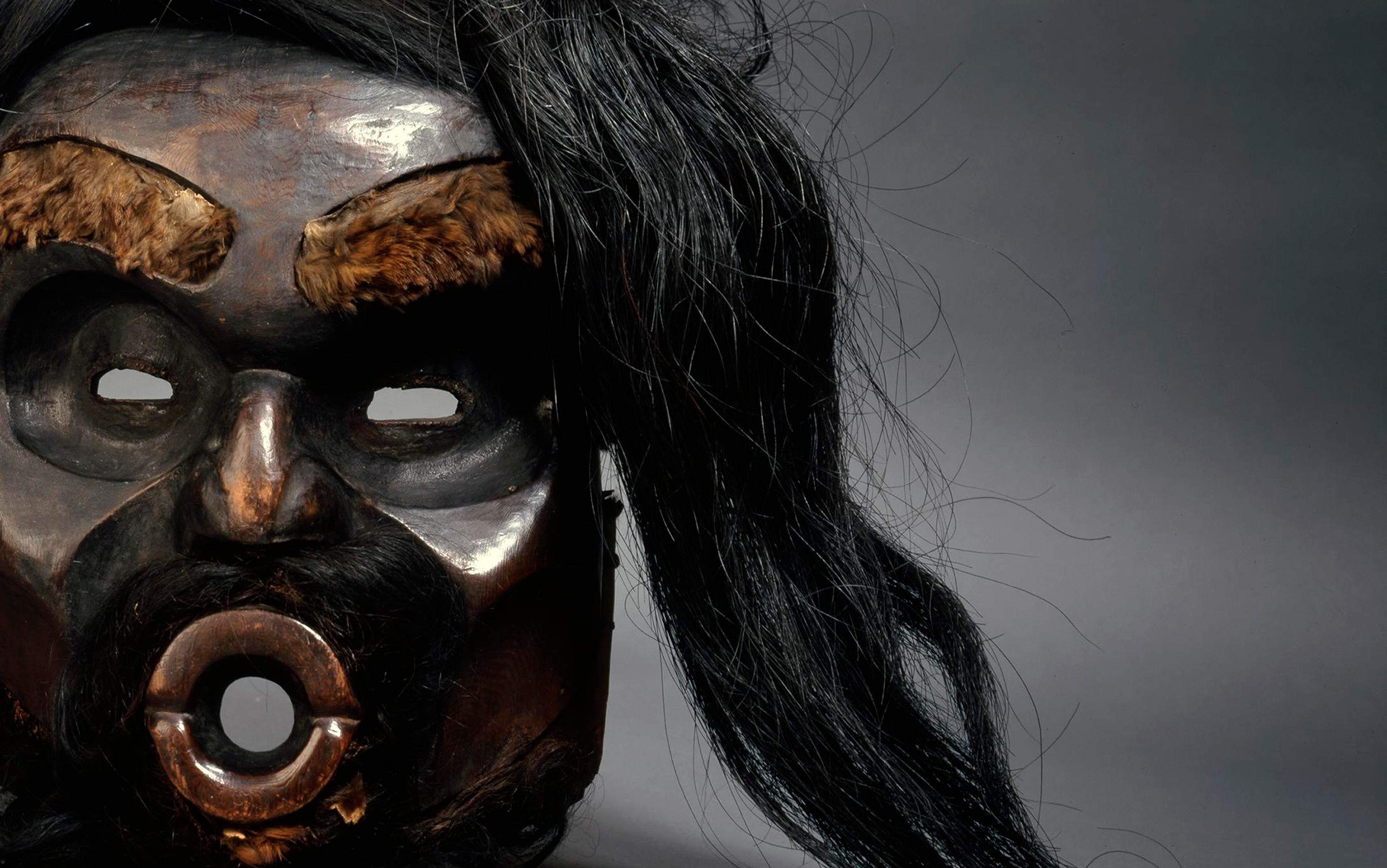 Close-up photo of a wooden mask with fur eyebrows, hollow eyes, a rounded mouth opening and long black hair against a dark background.