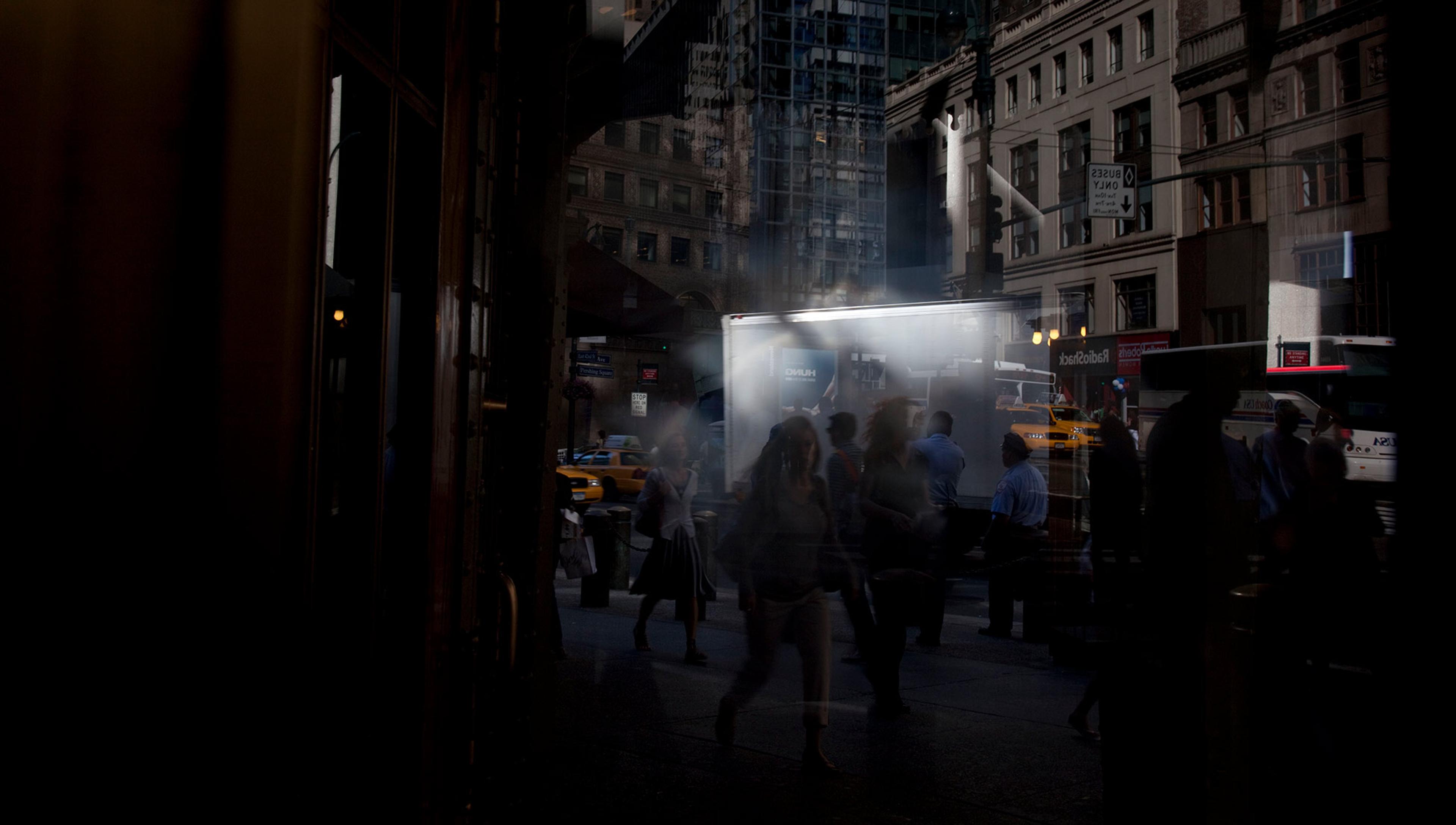 Busy city street at dusk with people walking, illuminated by bright lights from buildings and vehicles, including a lit-up truck and taxis in the background.