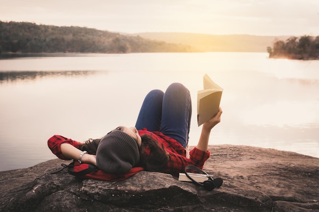 Foto mulher jovem deitada em uma rocha lendo um livro ao lado do lago contra um céu claro