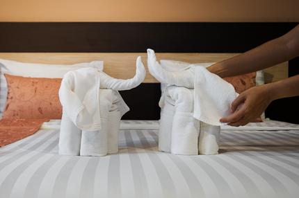 Berufe: Hotel maid cleaning bedroom service. Close-up of hands folding white bath towels  like elephant on the bed sheet.