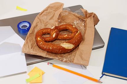 Langsam essen: A freshly baked pretzel snack on kraft paper amid office supplies, including a notebook, pencil, and adhesive notes.