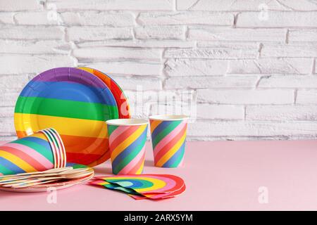 Rainbow colored disposable eco tableware on pink color background over white brick wall. Birthday party accessories,picnic utensil concept. Copy space Stock Photo