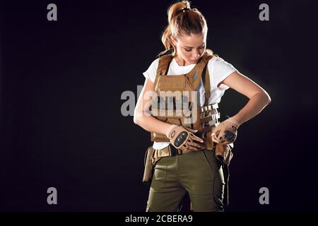 Gorgeous young woman with long blonde hair wearing Military gear, posing isolated in studio over dark background Stock Photo