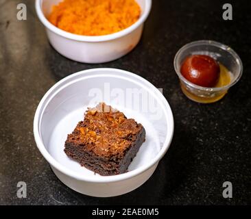 Chocolate Almond Brownie in a white takeout box on a black granite background. Gulab jamun and Carrot Halwa blurred in the background. Stock Photo