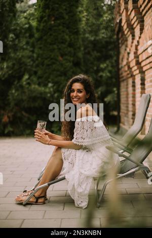 Pretty young woman with curly hair relaxes on a deck chair and enjoys a glass of fresh lemonade Stock Photo