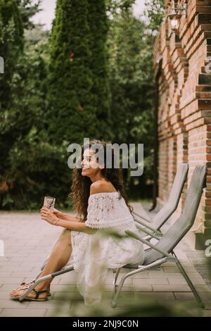 Pretty young woman with curly hair relaxes on a deck chair and enjoys a glass of fresh lemonade Stock Photo