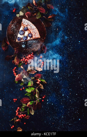 Floral border with a piece of blueberry pie, autumn leaves, berries and blue smoke on a dark wooden background. Halloween concept with copy space. Autumn flat lay with mystic smoke. Stock Photo