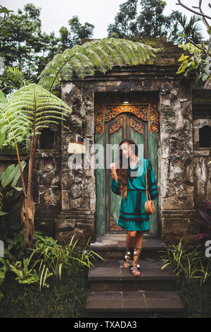 Asian brunette woman in green dress with handmade clutch bag posing on background of traditional balinese architecture Stock Photo