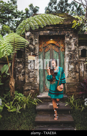 Asian brunette woman in green dress with handmade clutch bag posing on background of traditional balinese architecture Stock Photo