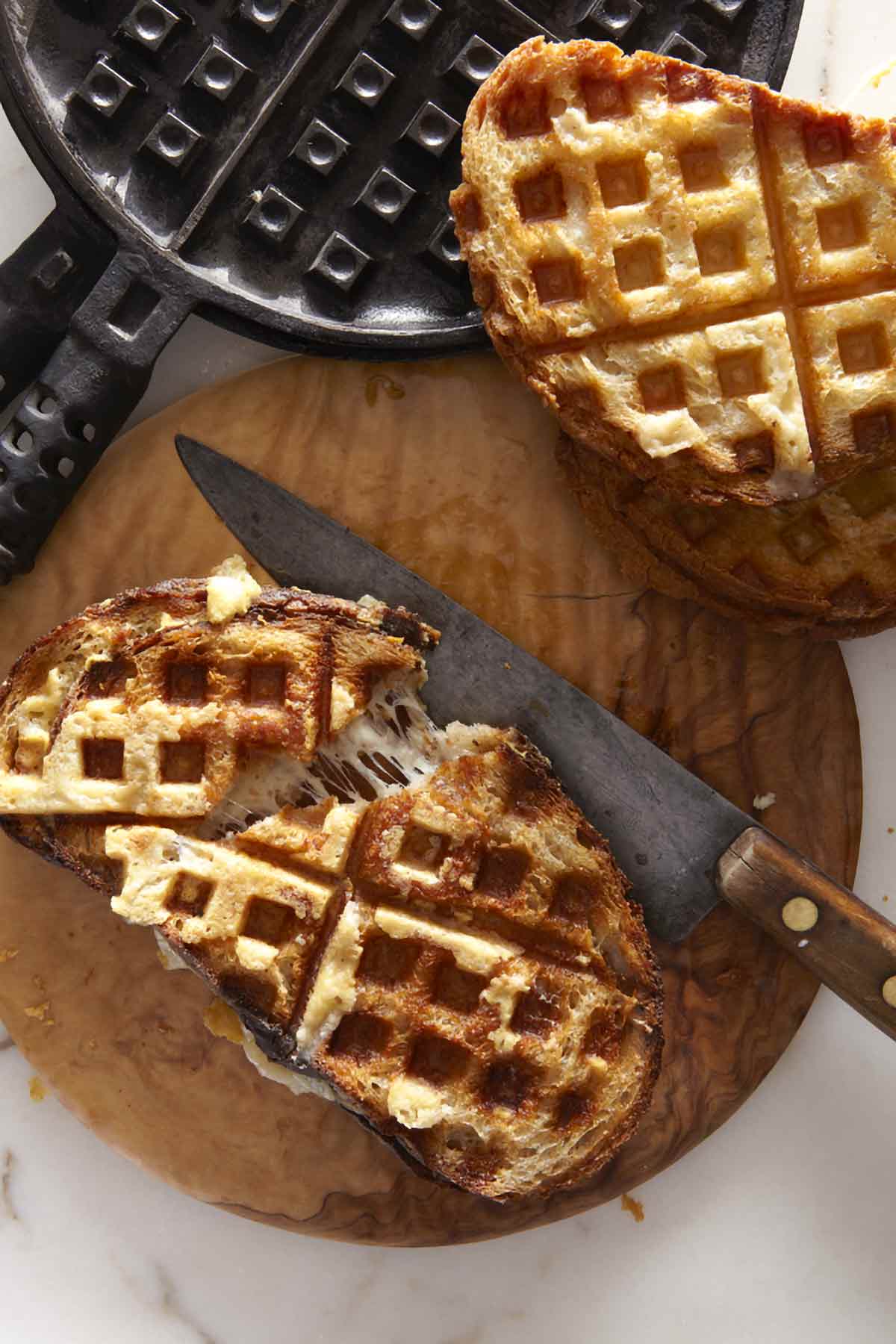 Two waffle iron grilled cheese sandwiches on a wooden cutting board with a knife and a waffle iron nearby.