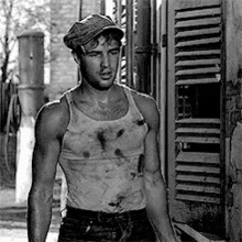 a man in a tank top and hat is standing in front of a building in a black and white photo .