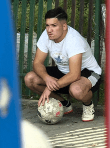 a man squatting down with a soccer ball wearing a white shirt that says converse