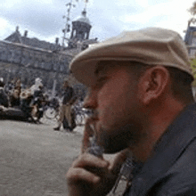 a man wearing a flat cap is smoking a cigarette in front of a crowd of people .