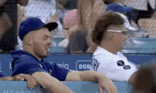 two men are sitting in the stands at a baseball game . one of the men is wearing a dodgers hat .