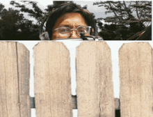 a man wearing headphones and glasses looks over a wooden fence