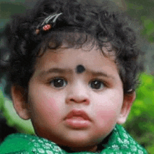 a baby girl with curly hair and a black spot on her forehead is wearing a green dress and looking at the camera .