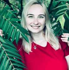 a woman wearing a red shirt and a heart shaped necklace smiles while holding green leaves