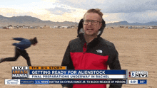 a man in a red and black north face jacket is standing in front of a screen that says getting ready for alienstock