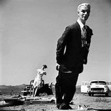 a man in a suit and tie is standing in the dirt in front of a car .