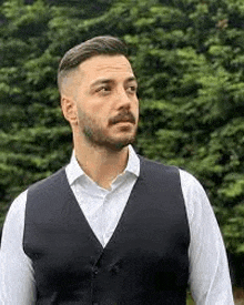 a man with a beard wearing a vest and a white shirt is standing in front of a forest .