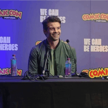 a man sits at a table in front of a sign that says we can be heroes