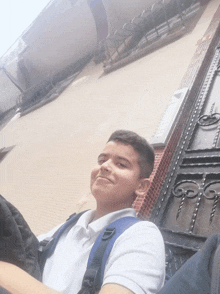 a boy wearing a white shirt and a blue backpack smiles in front of a building