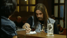 a man with long hair and a mustache is writing on a piece of paper next to a glass of beer