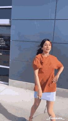 a woman in an orange shirt and blue skirt is dancing in front of a building that says united states postal service