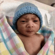 a baby wearing a blue hat is wrapped in a blanket and looking at the camera .