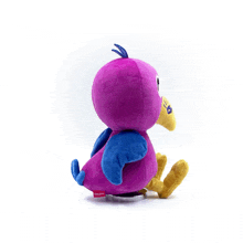 a stuffed purple bird with blue wings and a yellow beak is sitting on a white surface