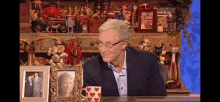 a man in a suit and glasses is sitting in front of a shelf with pictures and a mug with hearts on it ..