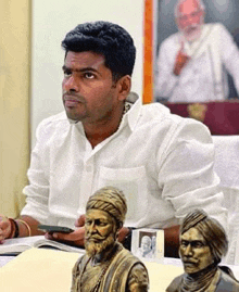 a man in a white shirt is sitting at a table with two statue of men .