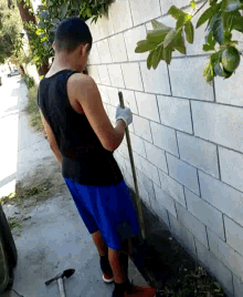 a man in a black tank top and blue shorts is holding a shovel in front of a brick wall