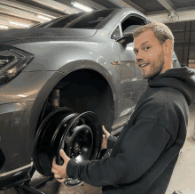 a man in a black hoodie is holding a black wheel in front of a car