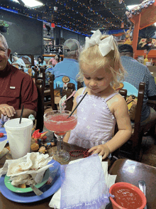 a little girl in a purple dress sits at a table with a margarita in front of her