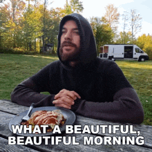 a man in a hoodie sits at a picnic table with a plate of food and the words what a beautiful beautiful morning above him