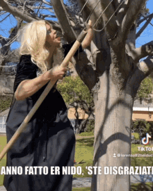 a woman in a black dress is holding a large wooden stick in front of a tree with the caption anno fatto er nido