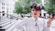 a woman wearing a white shirt and a black hat is standing in front of a building with the bbc logo on it